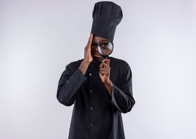 Young anxious afro-american cook in chef uniform looks through magnifying glass or loupe on white  with copy space