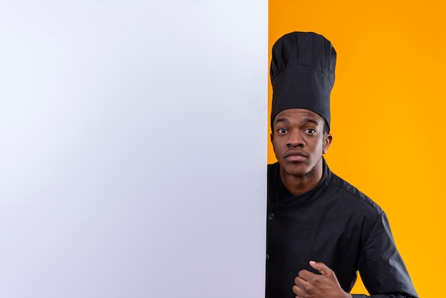 Молодой взволнованный афро-американский повар в униформе шеф-повара стоит за белой стеной и смотрит в камеру, изолированную на оранжевом фоне с копией пространства