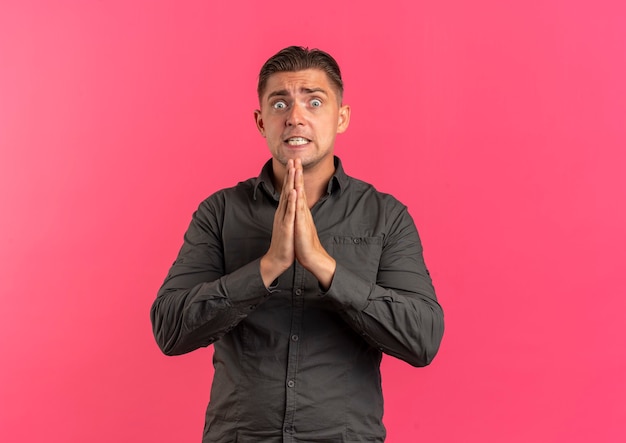 Young anxious blonde handsome man holds hands together isolated on pink background with copy space