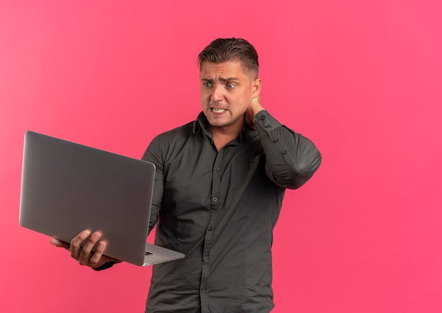 Young anxious blonde handsome man holds neck behind and looks at laptop isolated on pink background with copy space