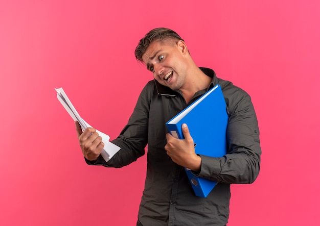 Young anxious blonde handsome man holds paper sheets and file folders talking on phone isolated on pink space with copy space