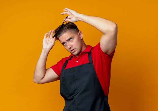 Young anxious blonde male barber in uniform combing hair and looks up isolated on orange space with copy space