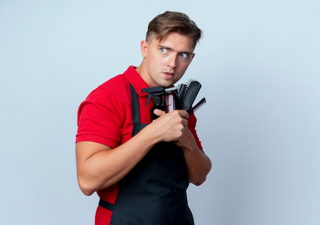 Free photo young anxious blonde male barber in uniform holds barber tools looking at side isolated on white space with copy space