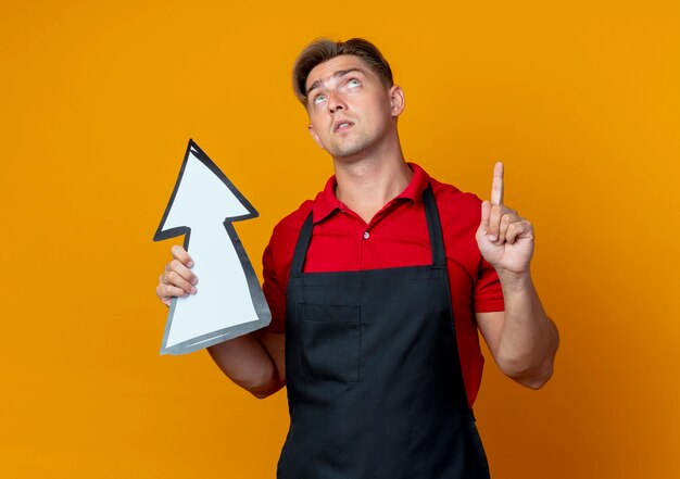 Young anxious blonde male barber in uniform holds direction mark looking and pointing up isolated on orange space with copy space