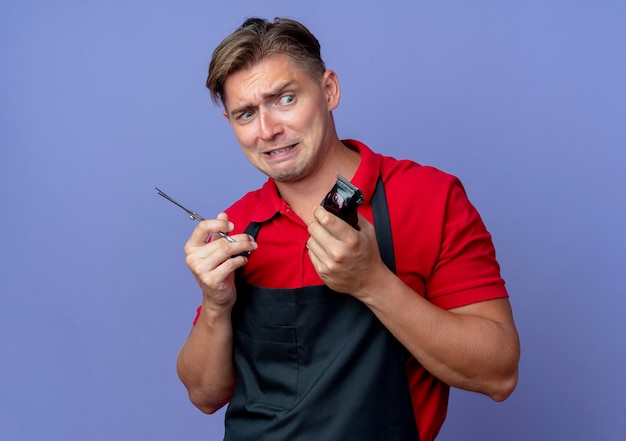 Young anxious blonde male barber in uniform holds scissors and hair clipper isolated on violet space with copy space