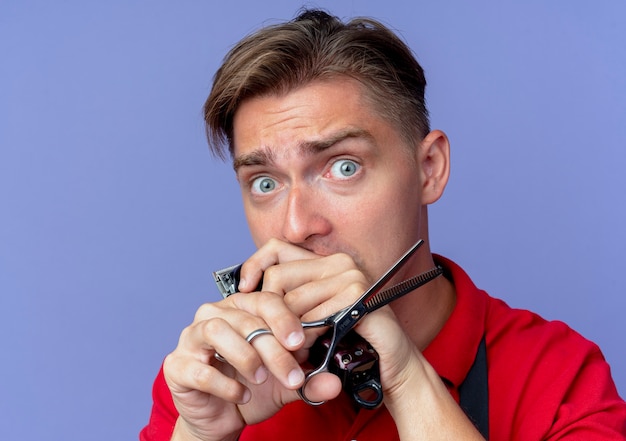 Young anxious blonde male barber in uniform holds scissors and hair clipper isolated on violet space with copy space