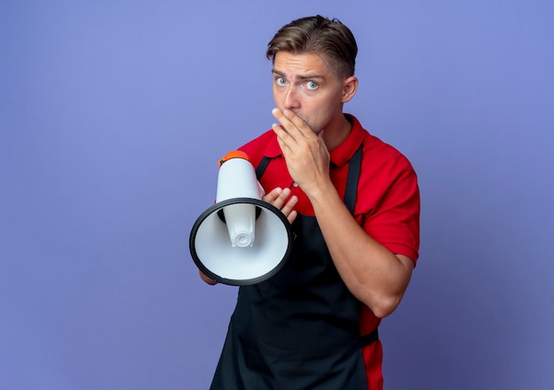Young anxious blonde male barber in uniform puts hand on mouth holding loud speaker isolated on violet space with copy space