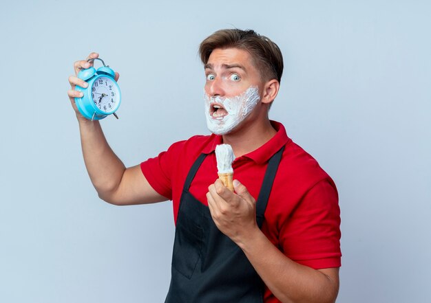 Young anxious blonde male barber in uniform smeared face with shaving foam holding alarm clock and shaving brush isolated on white space with copy space