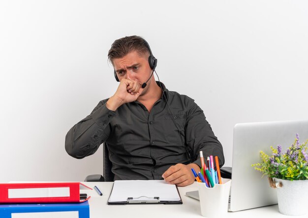 Young anxious blonde office worker man on headphones sits at desk with office tools using laptop bites fist looking down isolated on white background with copy space