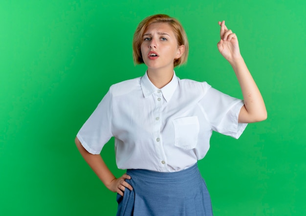 Free photo young anxious blonde russian girl crosses fingers looking at camera isolated on green background with copy space