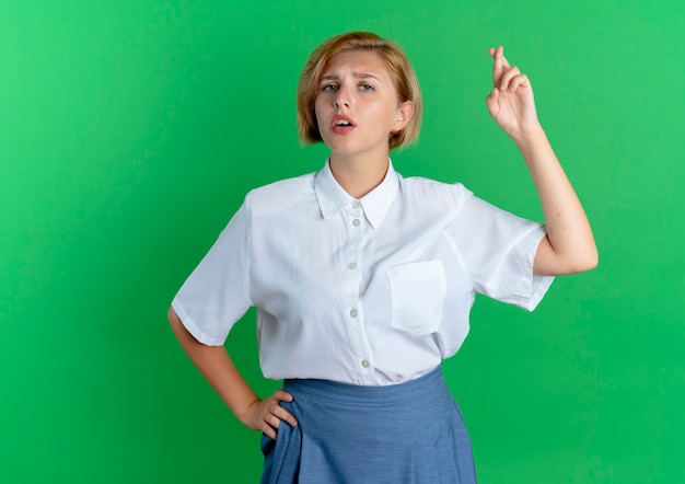 Young anxious blonde russian girl crosses fingers looking at camera isolated on green background with copy space