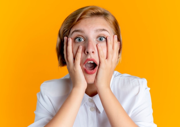 Young anxious blonde russian girl holds face looking at camera isolated on orange background with copy space