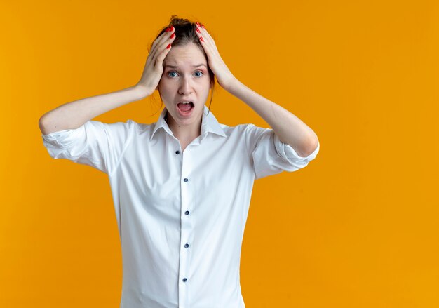 Young anxious blonde russian girl holds head isolated on orange space with copy space