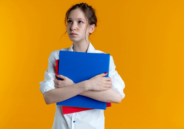 Young anxious blonde russian girl looks at side holding file folders on orange  with copy space