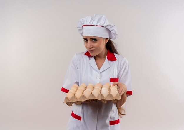 Young anxious caucasian cook girl in chef uniform holds batch of eggs and looks at camera isolated on white background with copy space