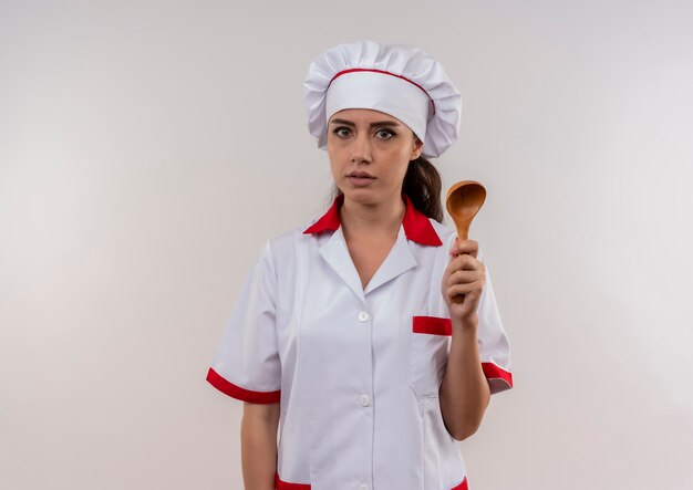 Young anxious caucasian cook girl in chef uniform holds wooden spoon isolated on white background with copy space