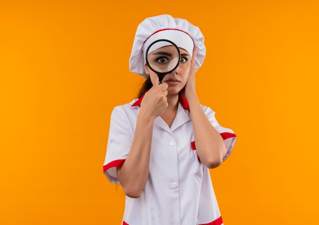 シェフの制服を着た若い気になる白人料理人の女の子は、コピースペースでオレンジ色の背景に分離された虫眼鏡またはルーペを通して見えます