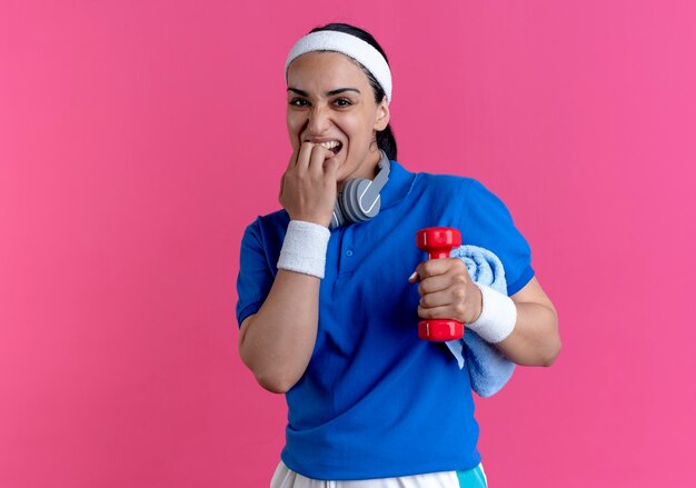 Молодая взволнованная кавказская спортивная женщина с головной повязкой и браслетами кусает пальцы, держа гантели и полотенце, изолированные на розовом фоне с копией пространства