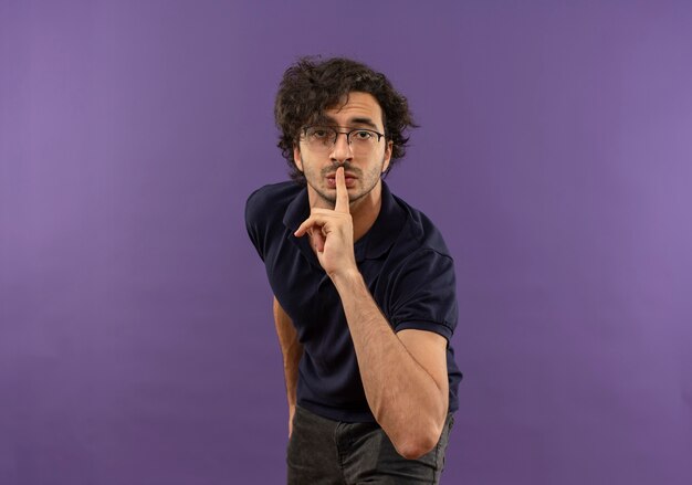 Young anxious man in black shirt with optical glasses gestures silence sign and looks isolated on violet wall