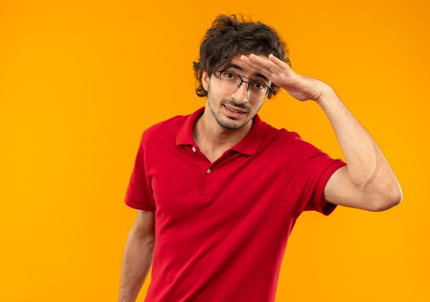 Young anxious man in red shirt with optical glasses keeps palm at forehead trying to see something distantly isolated on orange wall