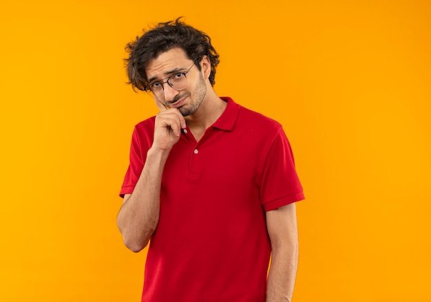 Молодой взволнованный мужчина в красной рубашке с оптическими очками кладет руку на подбородок и выглядит изолированным на оранжевой стене