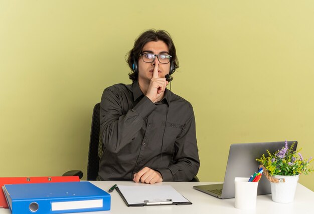 Молодой озабоченный офисный работник человек в наушниках в оптических очках сидит за столом с офисными инструментами, используя ноутбук, кладет палец в рот, жестикулируя тишину, изолированную на зеленом фоне с копией пространства
