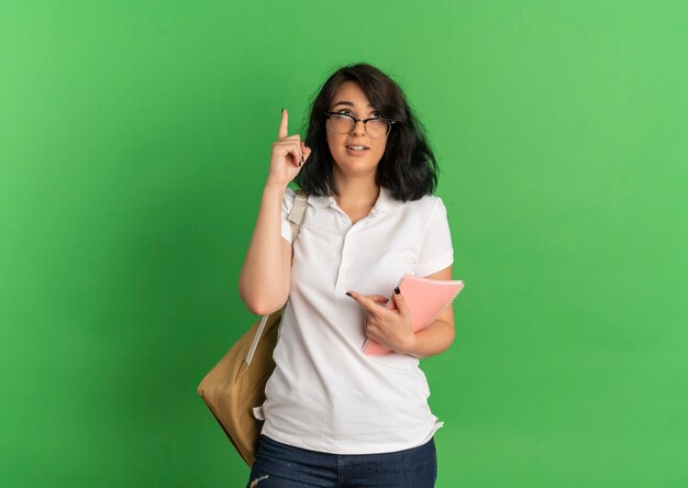 Young anxious pretty caucasian schoolgirl wearing glasses and back bag points and looks up holding notebook on green  with copy space