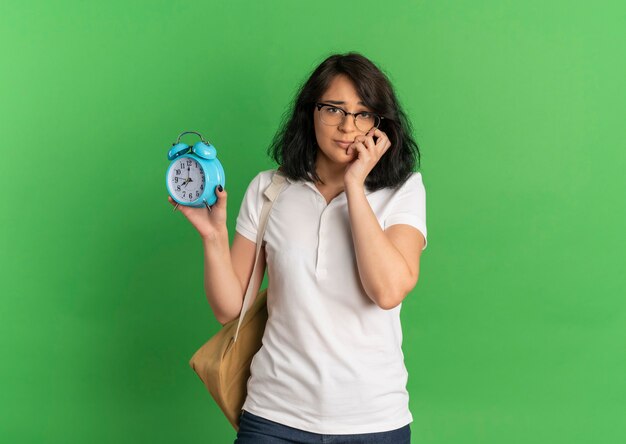 Young anxious pretty caucasian schoolgirl wearing glasses and back bag puts hand on face holding clock on green  with copy space