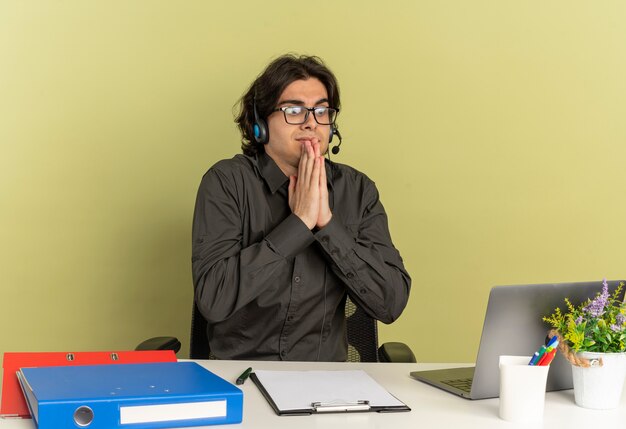 Молодой человек офисного работника в наушниках в оптических очках сидит за столом с офисными инструментами, используя и глядя на ноутбук, взявшись за руки вместе
