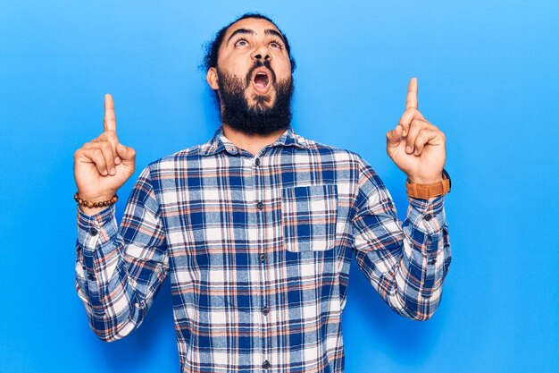 Young arab man wearing casual clothes amazed and surprised looking up and pointing with fingers and raised arms