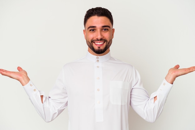 Young arab man wearing typical arab clothes isolated on white background makes scale with arms, feels happy and confident.