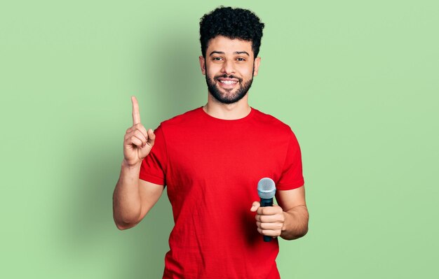 無料写真 マイクを使って歌を歌うひげを生やした若いアラブ人男性が、幸せそうな顔で指を差しながらアイデアや質問をしながら笑顔、ナンバーワン