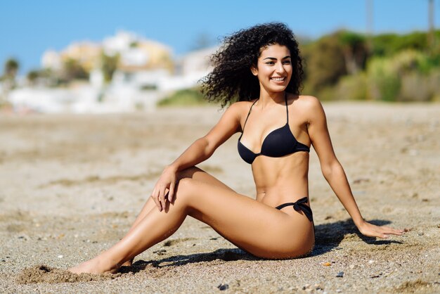 ビーチの砂に座っている水着で美しい体の若いアラビア人の女性。