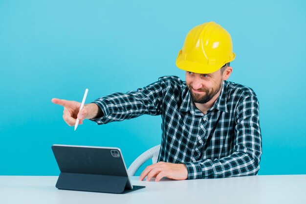 Young architect is pointing left with forefinger by sitting in front of tablet on blue background