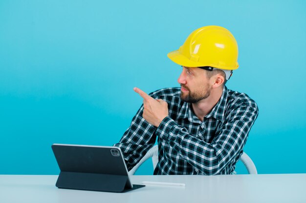 Young architect is pointing left with forefinger by sitting in front of tablet on blue background