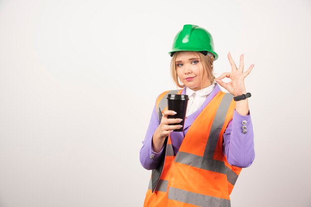 Young architect woman with clipboard and black cup showing ok gesture on white.