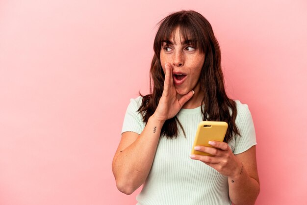 Молодая аргентинская женщина, держащая мобильный телефон на розовом фоне, говорит секретные горячие новости о торможении и смотрит в сторону