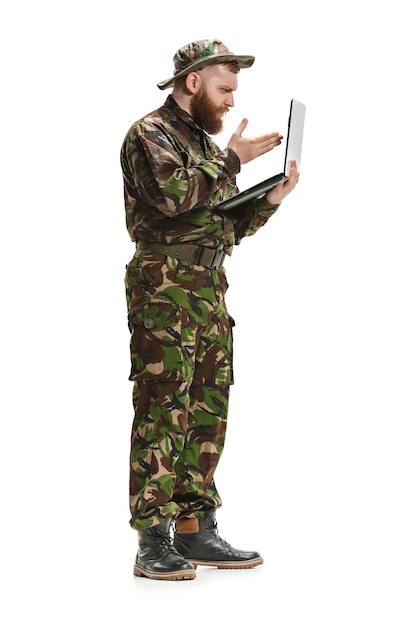 ノートパソコンとフルレングスの白いスタジオ背景に分離された迷彩服を着ている若い陸軍兵士。軍事、兵士、軍のコンセプトです。プロフェッショナル、コミュニケーション、コネクテッドピープルのコンセプト
