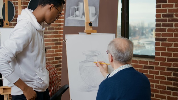 Young artist explaining drawing technique to elder person, using pencil to develop artistic skills on canvas and easel. Senior student learning to draw vase model in art class with teacher.