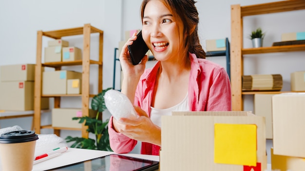 Free photo young asia businesswoman using mobile phone call receiving purchase order and check product on stock, work at home office. small business owner, online market delivery, lifestyle freelance concept.