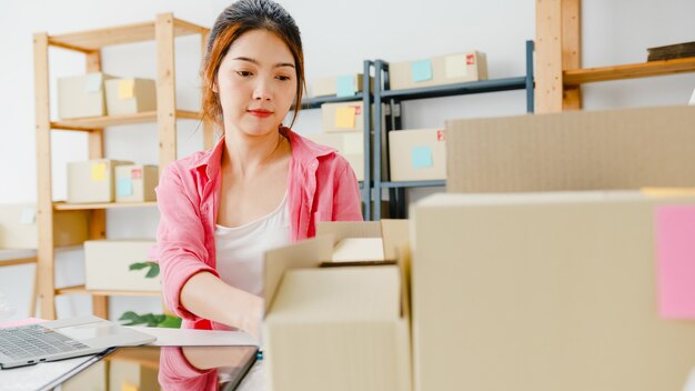 若いアジア起業家の実業家が在庫の製品購入注文をチェックし、ホームオフィスでコンピューターのラップトップの仕事に保存します。中小企業のオーナー、オンライン市場の配信、ライフスタイルのフリーランスのコンセプト。