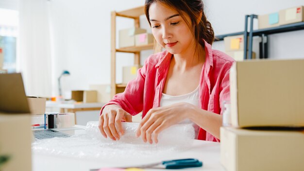 Young Asia entrepreneur businesswoman packing product in cardboard box deliver to customer, working at home office. Small business owner, start up online market delivery, lifestyle freelance concept.