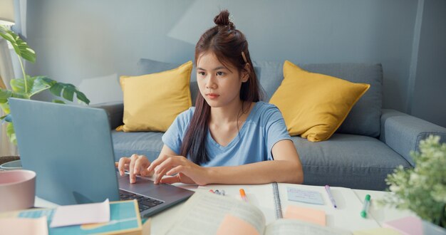 Молодая азиатская девушка-подросток с повседневным использованием портативного компьютера учится онлайн писать лекционную тетрадь для финального теста в гостиной дома