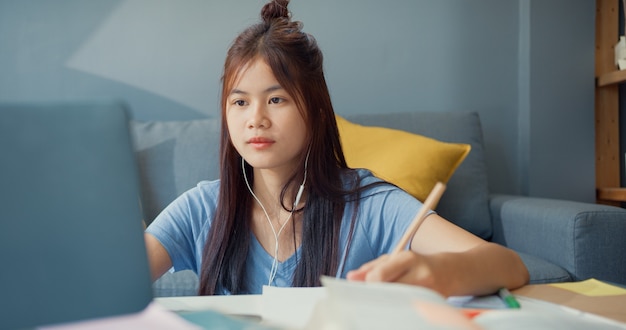 Молодая азиатская девушка-подросток с повседневной одеждой в наушниках использует портативный компьютер, учится онлайн писать лекционный блокнот в гостиной дома