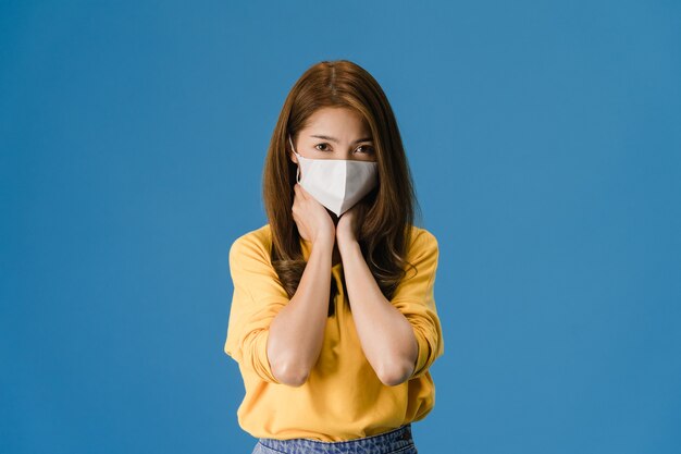 Молодая азиатская девушка в медицинской маске, уставшая от стресса и напряжения, уверенно смотрит в камеру, изолированную на синем фоне. Самоизоляция, социальное дистанцирование, карантин для предотвращения вируса короны.