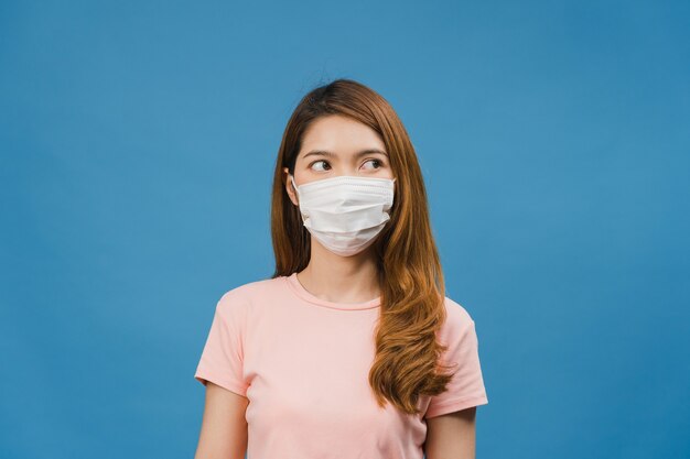 若いアジアの女の子は、ストレスと緊張にうんざりしている医療用フェイスマスクを着用し、青い壁に隔離されたスペースを自信を持って見ています