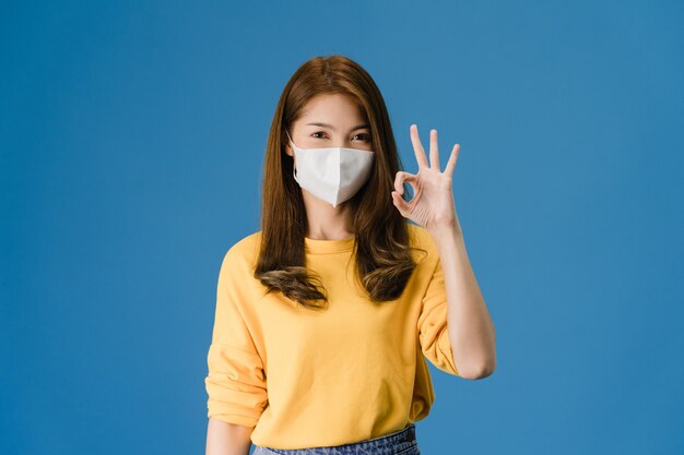 의료 얼굴 마스크를 착용하는 젊은 아시아 소녀 캐주얼 옷을 입고 확인 서명 몸짓과 파란색 배경에 고립 된 카메라를 봐. 자가 격리, 사회적 거리두기, 코로나 바이러스 격리.
