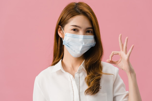 カジュアルな服を着て、ピンクの背景に分離されたカメラを見て、OKサインを身振りで示す医療フェイスマスクを身に着けている若いアジアの女の子。