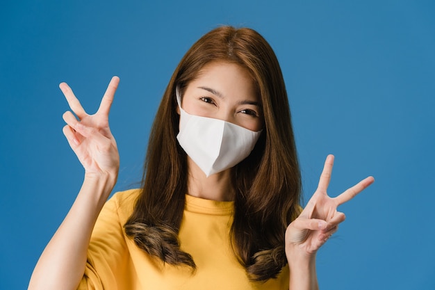 Young Asia girl wearing medical face mask showing peace sign, encourage with dressed in casual cloth and looking at camera isolated on blue background. Social distancing, quarantine for corona virus.