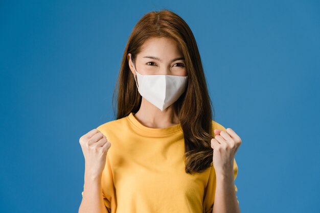 평화 기호를 보여주는 의료 얼굴 마스크를 착용하는 젊은 아시아 소녀, 캐주얼 옷을 입고 파란색 배경에 고립 된 카메라를보고 격려. 코로나 바이러스에 대한 사회적 거리두기, 격리.